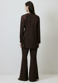 Costum două piese, maro, format dintr-un top cu mânecă lungă, cu spate plisat, și pantaloni evazați. Material neted cu detalii cu nasturi la gât.