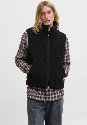Joven con el pelo largo y rubio, vistiendo un chaleco de forro polar negro con cremallera sobre una camisa de cuadros marrón y blanca y jeans oscuros.