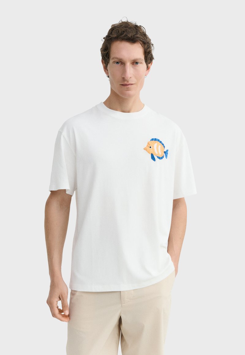 Homme portant un t-shirt blanc avec un poisson tropical coloré brodé sur le côté gauche de la poitrine et un pantalon beige, debout devant un fond clair.