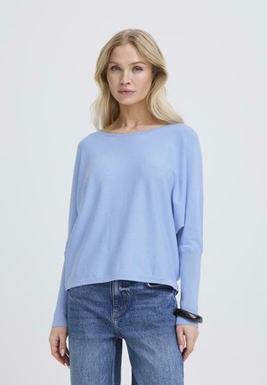 Donna bionda che indossa un maglione largo a maniche lunghe azzurro e jeans blu, con un braccialetto nero al polso destro, in piedi contro uno sfondo bianco.