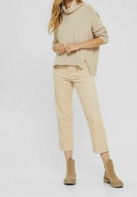 Kvinna klädd i en beige tröja, korta beige byxor och ljusbruna mocka stövlar som står framför en enfärgad bakgrund.