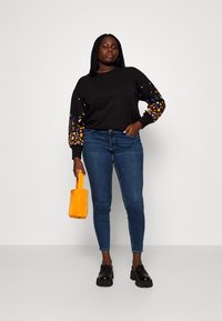 Sieviete, kas ģērbusies melnā džemperī ar ziedu raksta piedurknēm, zilos pieguļošos džinsos, melnās masīvās mokasīnēs un rokās tur nelielu dzeltenu somiņu.