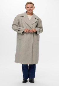 Jasnoszary, oversize'owy płaszcz z wełny z klapami, z przednimi kieszeniami i zapinanymi mankietami, noszony na kremowym swetrze i dżinsach.