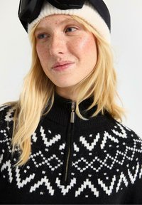 Sort zip-up sweater med hvide geometriske strikkemønstre. Høj krave og lynlåsdetalje. Blød tekstureret stof. Casual vintertøj.