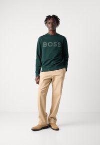 Felpa verde con logo "BOSS", abbinata a pantaloni beige chiaro. Il modello si trova su uno sfondo neutro, evidenziando i dettagli dell'abbigliamento.