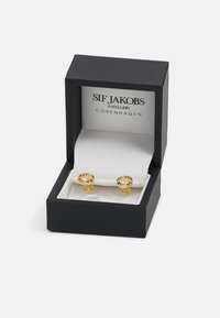 Guldörhängen med klara och bärnstensfärgade stenar, förvarade i en svart smyckeslåda med vit inredning och varumärkeslogotypen tydligt visad.