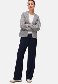 Femme debout, les bras croisés, portant un cardigan en tricot gris, une chemise blanche, un pantalon large bleu marine et des baskets blanches sur un fond uni.
