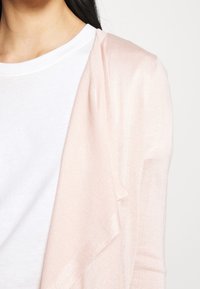 Světle růžový kardigan z měkké texturované látky. Drapovaný design má otevřený přední díl a mírně prodloužené rukávy, vrstvený přes bílou košili.