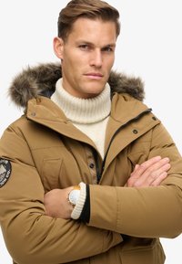 Chaqueta inflable marrón con capucha de piel sintética, con cremallera y botones a presión, combinada con un cuello alto de canalé en color crema y un reloj elegante.