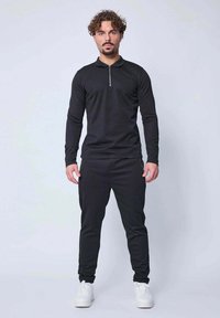 Haut noir à manches longues avec col zippé, associé à un pantalon jogger noir. Tissu lisse et design ajusté, complété par des baskets blanches.