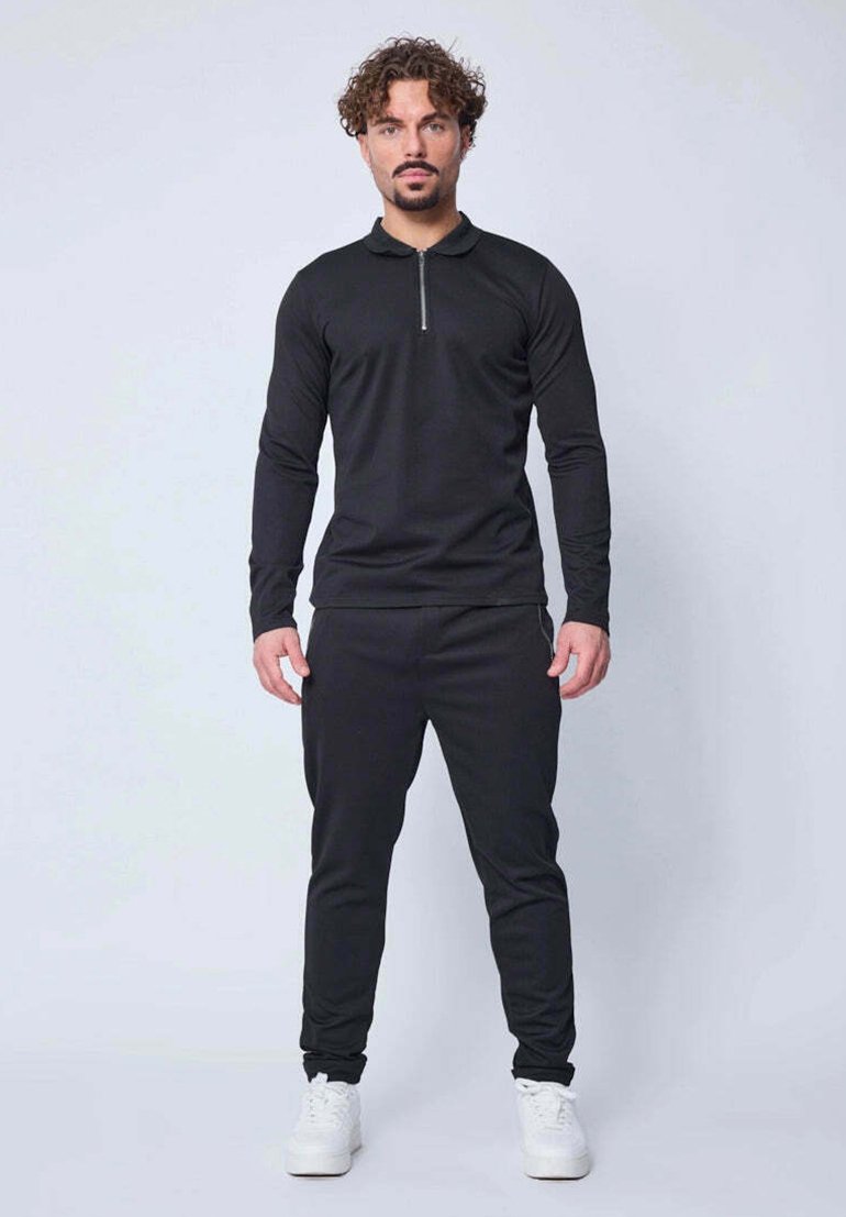 Haut noir à manches longues avec col zippé, associé à un pantalon jogger noir. Tissu lisse et design ajusté, complété par des baskets blanches.