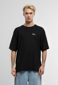 Černé oversized tričko z bavlny s kulatým výstřihem, krátkými rukávy a malým bílým logem na levé části hrudi. Hladká textura.