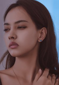 Orecchini a forma di cuore in oro con accento di gemma blu indossati sulla orecchio di un modello, che mettono in mostra una finitura liscia e un design elegante.