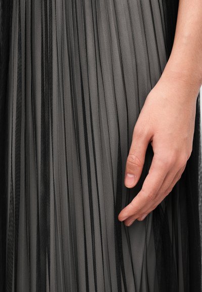 Μαύρο και γκρι πλισέ ύφασμα με διαφανή υφή, με κάθετες ρίγες. Ένα χέρι ακουμπά ελαφρά πάνω στο ύφασμα, επιδεικνύοντας τις στρώσεις του.
