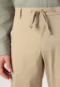 Beige bukser med elastisk talje og snor, med en glat tekstur og sidelommer. Kombineret med en grøn skjorte.