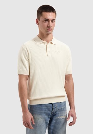 Junger Mann mit kurzem Haar, der ein cremefarbenes, strukturiertes Poloshirt mit kurzen Ärmeln und zerrissene blaue Jeans trägt, vor einem schlichten Hintergrund steht.