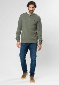 Bajuszós férfi olajzöld, hosszú ujjú henley pólót, kék farmert és világosbarna sportcipőt visel, egy egyszínű, világos háttér előtt áll.