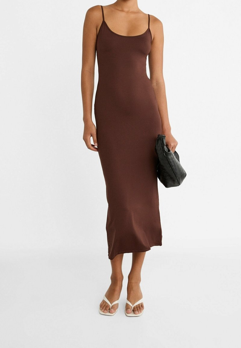 Femme portant une robe midi marron sans manches, tenant un sac à main noir tressé et portant des sandales blanches sur un fond blanc.