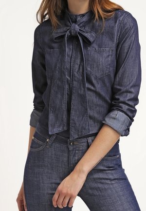 Femme portant une chemise en denim foncé avec un nœud noué au col et un jean en denim foncé assorti avec des manches retroussées.