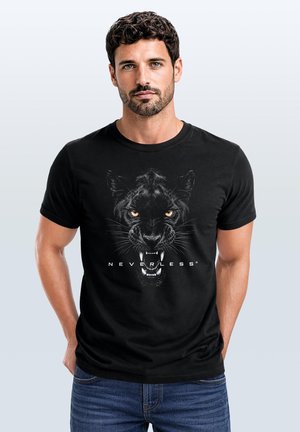 Mann mit dunklen lockigen Haaren und Bart, der ein schwarzes T-Shirt trägt, das ein fauchendes schwarzes Panthergesicht und das Wort "NEVERLESS" auf der Vorderseite zeigt.