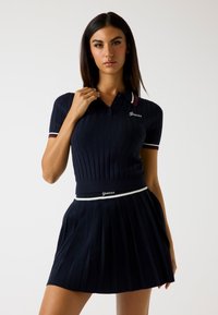 Polo a righe blu navy con maniche corte e colletto a contrasto, abbinato a una gonna plissettata con vita bianca e logo.