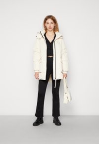 Dik wit jack met een capuchon, ritssluiting en elastische manchetten, gecombineerd met zwarte flared broek en stevige zwarte schoenen, met een witte tas in de hand.