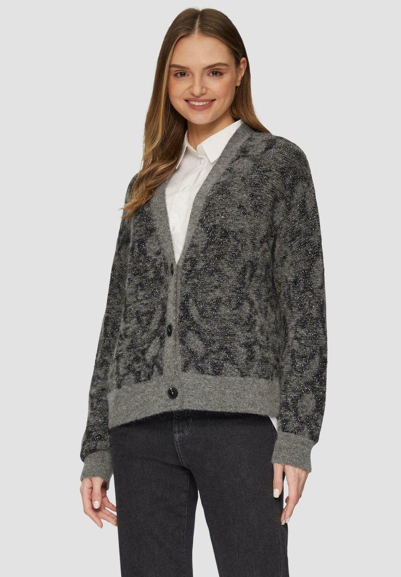 Grijze cardigan met een gestructureerd, sprankelend oppervlak en een knoopsluiting. Het heeft een V-hals en gebreide manchetten, gedragen over een witte blouse.