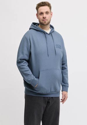 Mann med krøllete hår iført blå Jack & Jones-hettegenser og svarte bukser, stående med en hånd i lommen mot en ensfarget bakgrunn.
