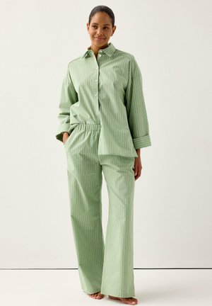 Kvinde står barfodet iført lysegrøn skjorte med fine striber og matchende bukser med vide ben, hænderne i lommerne, neutral baggrund.
