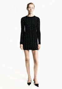 Abito nero lavorato a maglia con maniche lunghe, caratterizzato da un motivo testurizzato e piccoli dettagli metallici. Modello abbinato a décolleté nere a punta.