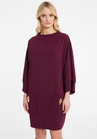 Weinrotes Kleid mit hohem Ausschnitt und weiten Ärmeln. Mit zwei vorderen Taschen und dekorativen goldenen Akzenten. Glatte Stoffstruktur.