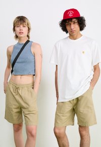 Blue ribbed crop top with black strap, beige drawstring shorts. Male model in white t-shirt with logo, each wearing matching beige shorts.