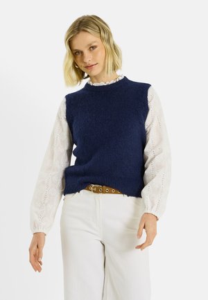 Mujer rubia con un chaleco sin mangas de punto azul marino sobre una blusa blanca de manga larga con estampado, pantalones blancos y un cinturón marrón, posando contra un fondo blanco.