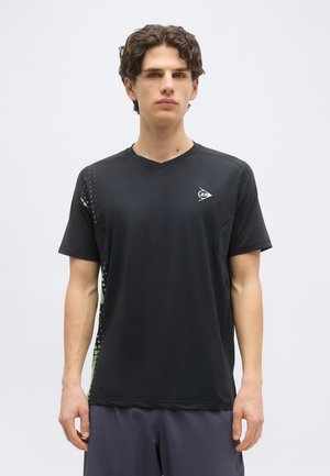 Νέος άνδρας με κατσαρά μαλλιά που φορεί μαύρο αθλητικό μπλουζάκι με λογότυπο στο στήθος και πλευρικά πάνελ με σχέδιο, στέκεται μπροστά σε απλό φόντο.