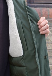 Grüne Jacke mit weichem, cremefarbenem Fleece-Futter, Seitentasche und sichtbaren Reißverschlussdetails. Glatter Außenstoff mit wattierter Polsterung.