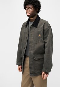 Νεαρό άτομο με λεύκη φοράει σκούρο πράσινο μπουφάν με μαύρο γιακά, γκρι πουκάμισο και μπεζ παντελόνι, στέκεται μπροστά σε απλό φόντο.