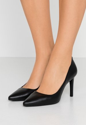 Femme portant des chaussures à talons hauts noires à bouts pointus, debout sur un sol lisse de couleur claire, devant un fond uni.