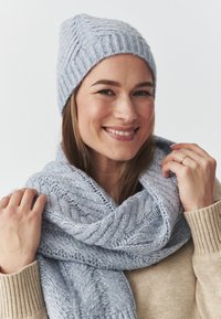 Căciulă și eșarfă tricotate de culoare albastru deschis, cu un model texturat, asortate cu un pulover bej. Eșarfa este aruncată lejer în jurul gâtului.