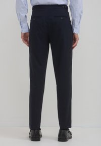 Marineblaue Tailored Trousers mit schmaler Passform, ausgestattet mit zwei Gesäßtaschen und einer glatten Textur, kombiniert mit schwarzen Halbschuhen.