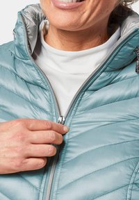 Giacca puffa azzurro chiaro con cuciture a chevron, colletto alto e zip frontale. Una mano sta regolando il tirazip.