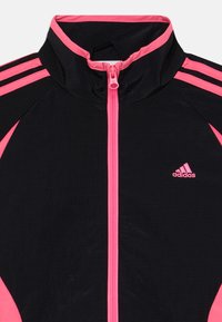 Jacket atlético preto com gola alta e detalhes em rosa, incluindo um zíper central e três riscas rosas em cada ombro.