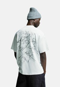 Man in grijs gebreide beanie, witte oversized T-shirt met abstracte zwarte lijntekening op de rug, en zwarte shorts, zijwaarts tegen een witte achtergrond.