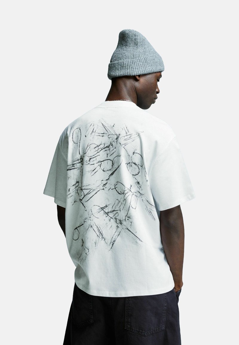 Man in grijs gebreide beanie, witte oversized T-shirt met abstracte zwarte lijntekening op de rug, en zwarte shorts, zijwaarts tegen een witte achtergrond.