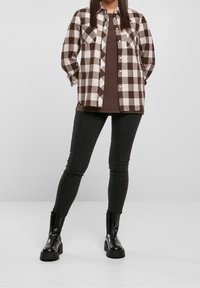 Camicia di flanella a scacchi marrone e bianca con due tasche frontali sopra una maglietta marrone, abbinata a jeans skinny neri e stivaletti neri.