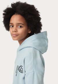 Felpa blu chiaro con una texture morbida, dotata di una tasca frontale e un logo grafico nero sulla manica sinistra. Design con cappuccio e orlo a coste.