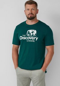 Dunkelgrünes Baumwoll-T-Shirt mit einem weißen Grafiklogo des Discovery Channel. Kurze Ärmel, Rundhalsausschnitt.