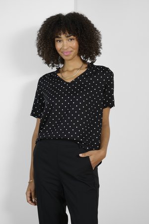 Zwarte blouse met polka dots, een v-hals, korte mouwen en een zachte textuur. Gecombineerd met hoge zwarte broeken met zijzakken.