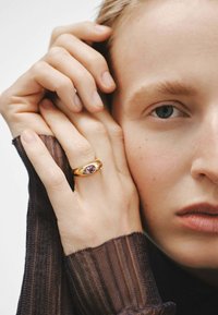 Anillo de oro con una gema de amatista ovalada, llevado en una mano con dedos delgados. La mano descansa sobre un fondo neutro.