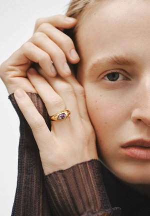 Anillo de oro con una gema de amatista ovalada, llevado en una mano con dedos delgados. La mano descansa sobre un fondo neutro.