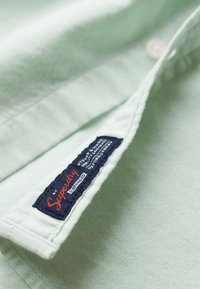 Lichtgroen hemd van gestructureerde stof, met een donkerblauw label met rode letters bij de kraag. Witte knoop zichtbaar.